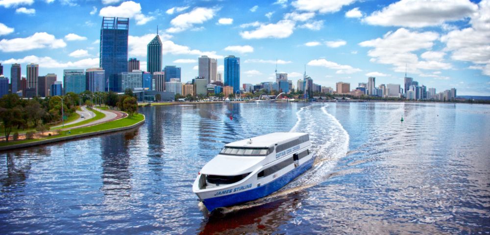 A white ferry glides along a shimmering river, with Perth's skyline and blue sky dotted with clouds in the background.