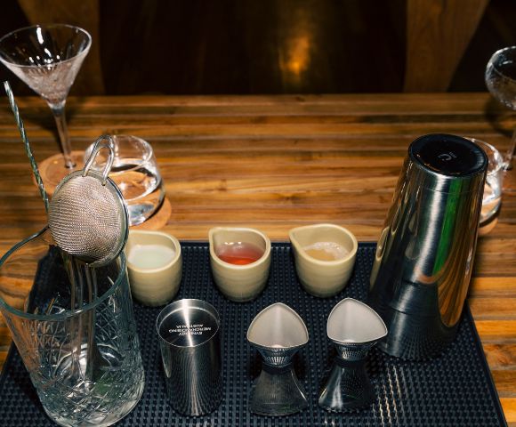 Cocktail bar setup with mixing glass, strainer, jigger, shaker, and small pitchers on a textured mat. The wooden table creates a warm, inviting atmosphere.