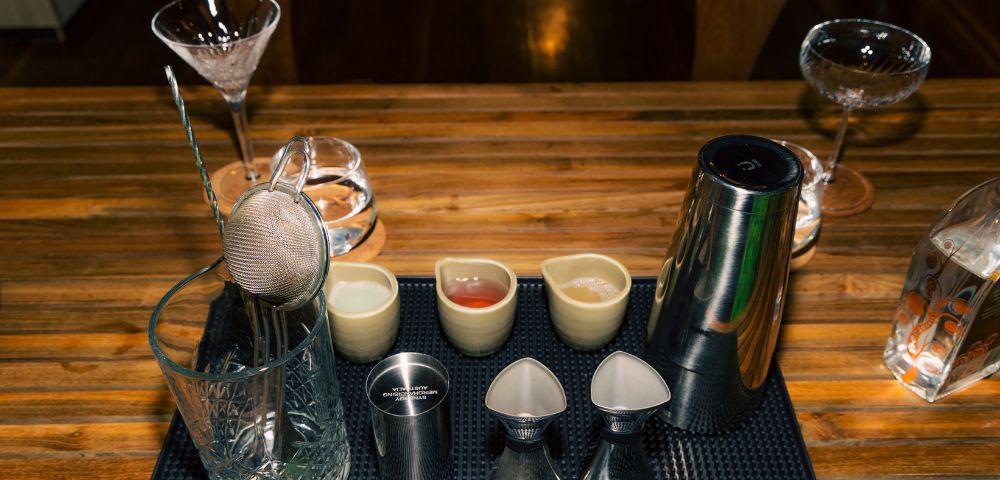 Cocktail bar setup with mixing glass, strainer, jigger, shaker, and small pitchers on a textured mat. The wooden table creates a warm, inviting atmosphere.