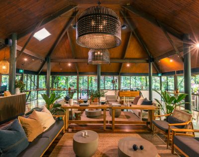 A cozy, rustic lounge with wooden ceilings and large wicker chandeliers. Stylish furniture and lush greenery create a warm, inviting atmosphere.