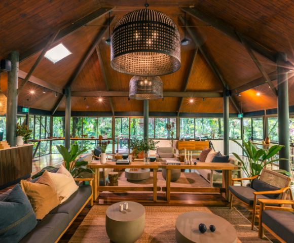 A cozy, rustic lounge with wooden ceilings and large wicker chandeliers. Stylish furniture and lush greenery create a warm, inviting atmosphere.
