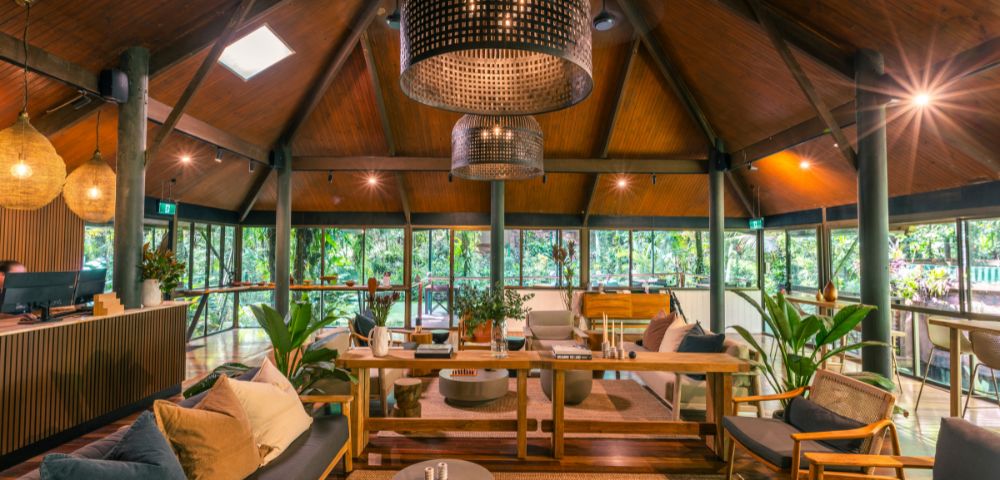 A cozy, rustic lounge with wooden ceilings and large wicker chandeliers. Stylish furniture and lush greenery create a warm, inviting atmosphere.