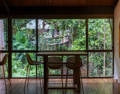A cozy indoor dining area with wooden floors and a table with three chairs facing a large window. Lush green forest scenery is visible outside.