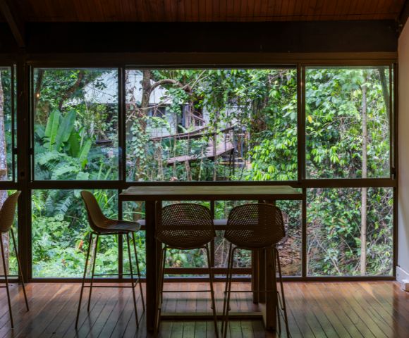 A cozy indoor dining area with wooden floors and a table with three chairs facing a large window. Lush green forest scenery is visible outside.