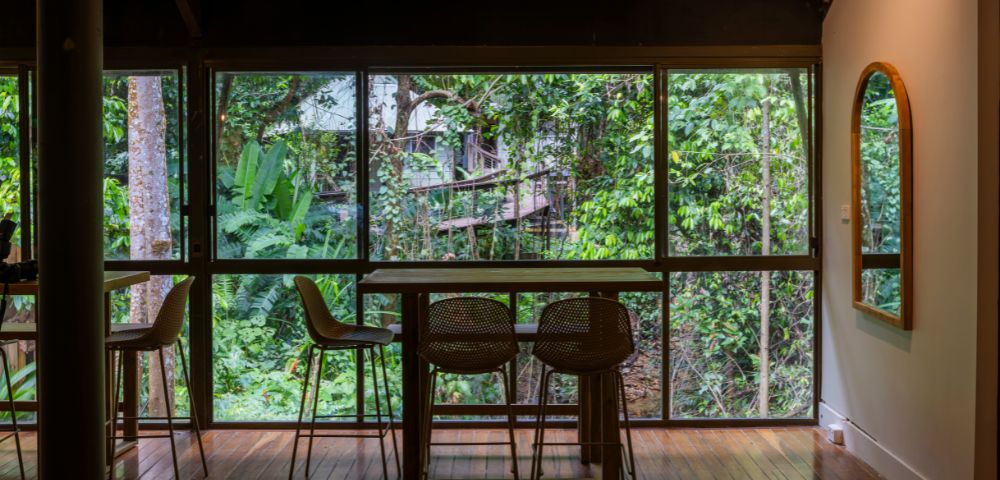 A cozy indoor dining area with wooden floors and a table with three chairs facing a large window. Lush green forest scenery is visible outside.