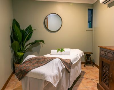 A serene massage room with a massage table covered in white linens and a patterned blanket. There’s a potted plant, a round mirror, and soft lighting.