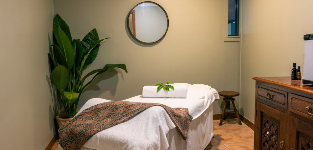 A serene massage room with a massage table covered in white linens and a patterned blanket. There’s a potted plant, a round mirror, and soft lighting.