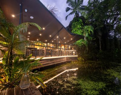 A modern restaurant with large windows and warm lighting sits amidst lush greenery by a tranquil pond at dusk, creating a serene atmosphere.