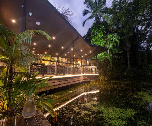 A modern restaurant with large windows and warm lighting sits amidst lush greenery by a tranquil pond at dusk, creating a serene atmosphere.