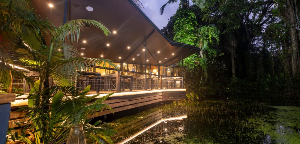 A modern restaurant with large windows and warm lighting sits amidst lush greenery by a tranquil pond at dusk, creating a serene atmosphere.