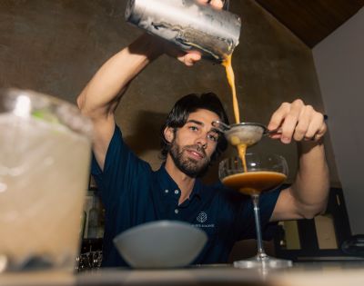 A bartender wearing a navy shirt is skillfully pouring a brown cocktail through a strainer into a coupe glass. The setting is a dimly lit bar.