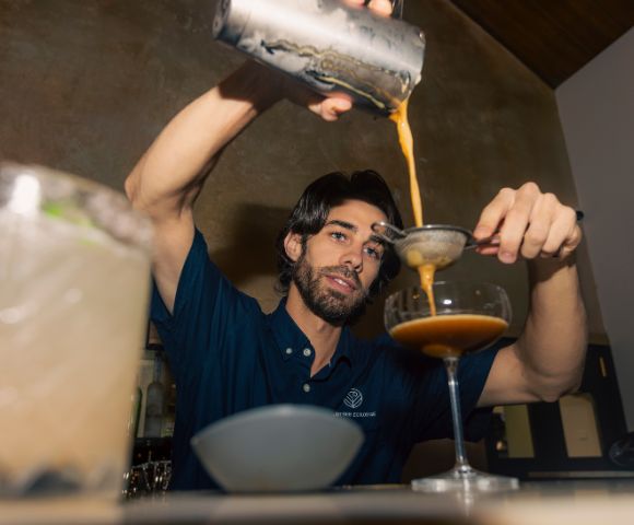 A bartender wearing a navy shirt is skillfully pouring a brown cocktail through a strainer into a coupe glass. The setting is a dimly lit bar.