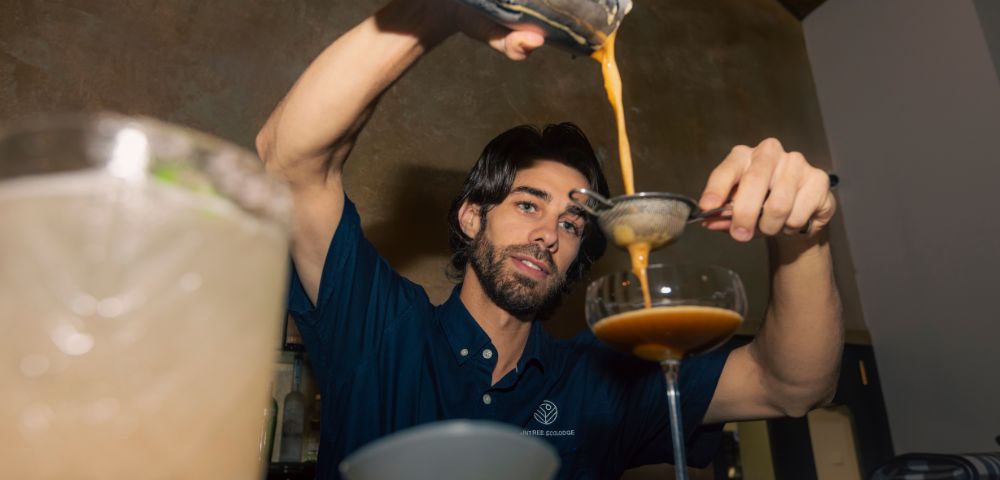 A bartender wearing a navy shirt is skillfully pouring a brown cocktail through a strainer into a coupe glass. The setting is a dimly lit bar.