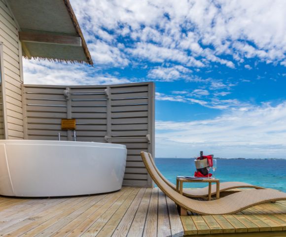 Luxurious deck overlooking the ocean with a white freestanding bathtub, wicker lounge chairs, and a table holding wine and glasses. Sky is partly cloudy.