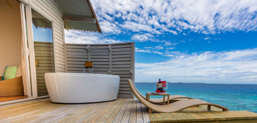 Luxurious deck overlooking the ocean with a white freestanding bathtub, wicker lounge chairs, and a table holding wine and glasses. Sky is partly cloudy.