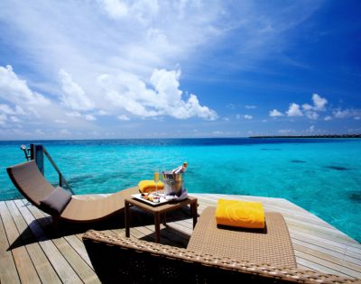 Sunlit deck overlooking turquoise ocean with two lounge chairs, yellow towels, and a table with champagne. Bright sky and tranquil ambiance.