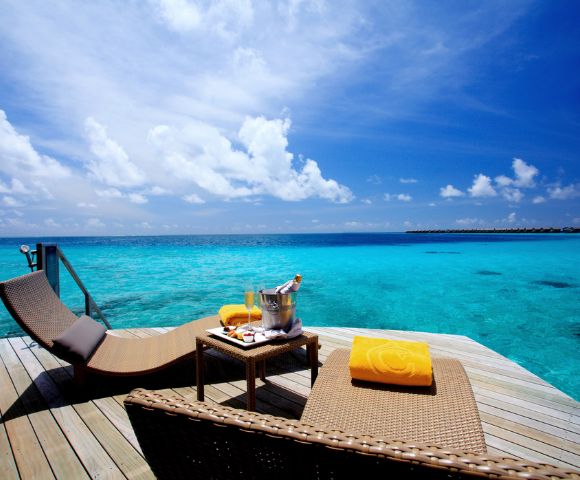 Sunlit deck overlooking turquoise ocean with two lounge chairs, yellow towels, and a table with champagne. Bright sky and tranquil ambiance.