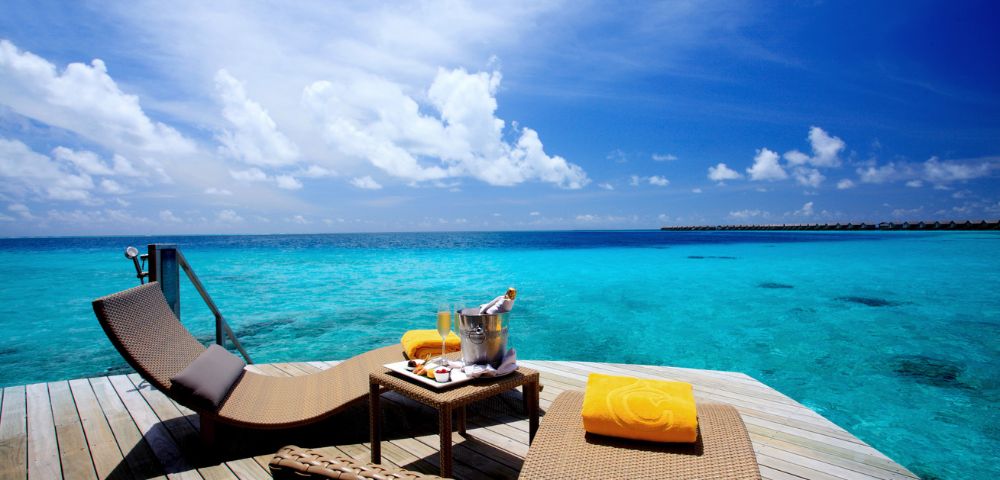 Sunlit deck overlooking turquoise ocean with two lounge chairs, yellow towels, and a table with champagne. Bright sky and tranquil ambiance.