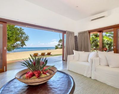 Spacious beachfront living room with open sliding doors reveals a sandy beach and ocean view. White sofas, tropical flower centerpiece, relaxed vibe.