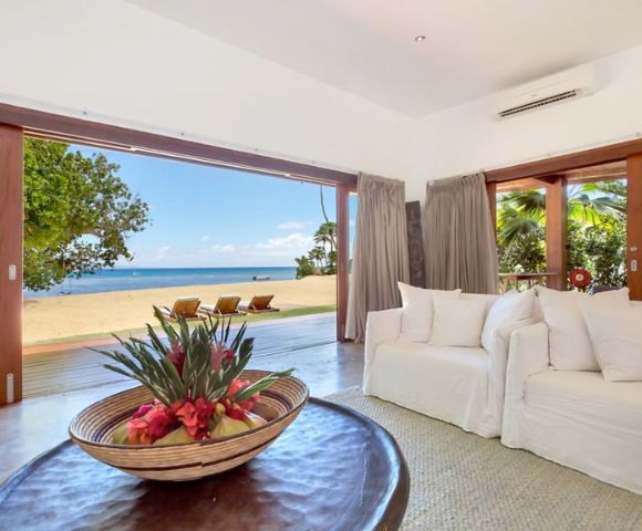 Spacious beachfront living room with open sliding doors reveals a sandy beach and ocean view. White sofas, tropical flower centerpiece, relaxed vibe.