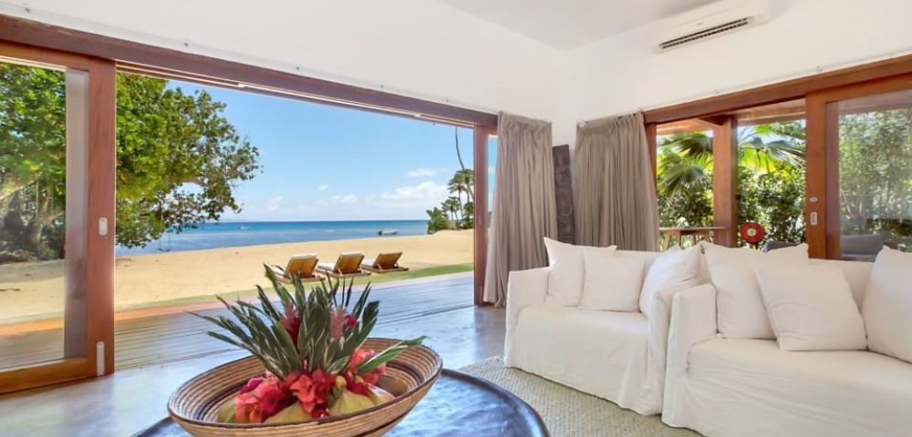 Spacious beachfront living room with open sliding doors reveals a sandy beach and ocean view. White sofas, tropical flower centerpiece, relaxed vibe.