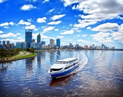 A white ferry travels across a calm river reflecting the vibrant blue sky and fluffy clouds, with a city skyline and green park visible in the background.