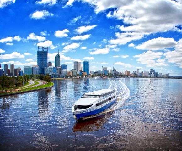 A white ferry travels across a calm river reflecting the vibrant blue sky and fluffy clouds, with a city skyline and green park visible in the background.