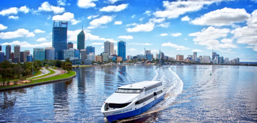 A white ferry travels across a calm river reflecting the vibrant blue sky and fluffy clouds, with a city skyline and green park visible in the background.