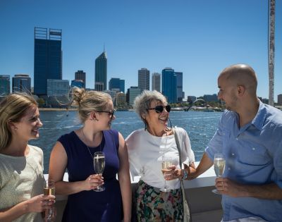 Four people, smiling and holding champagne glasses, stand on a boat with a city skyline in the background. The mood is joyful and celebratory.