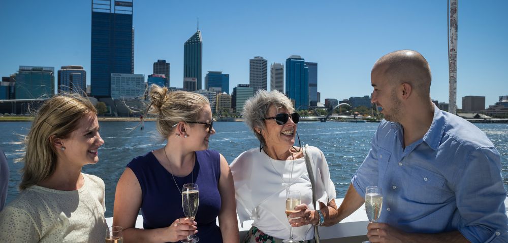 Four people, smiling and holding champagne glasses, stand on a boat with a city skyline in the background. The mood is joyful and celebratory.