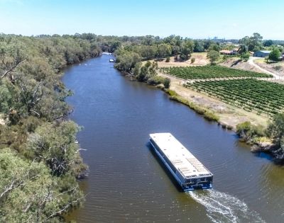 Aerial view of a river with a boat traveling through. Lush trees line the riverbanks, and a vineyard is visible on the right. The scene is peaceful and sunny.