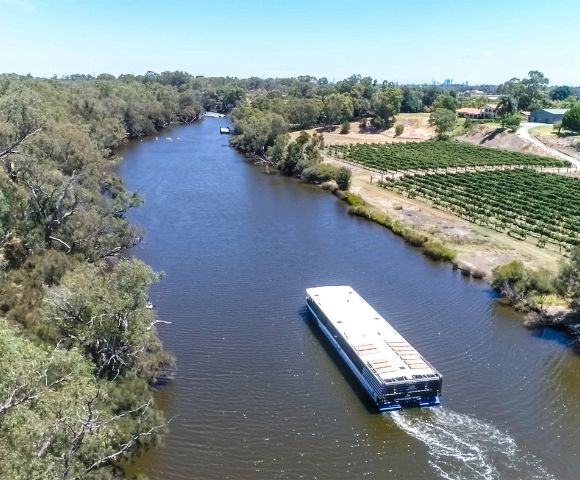 Aerial view of a river with a boat traveling through. Lush trees line the riverbanks, and a vineyard is visible on the right. The scene is peaceful and sunny.