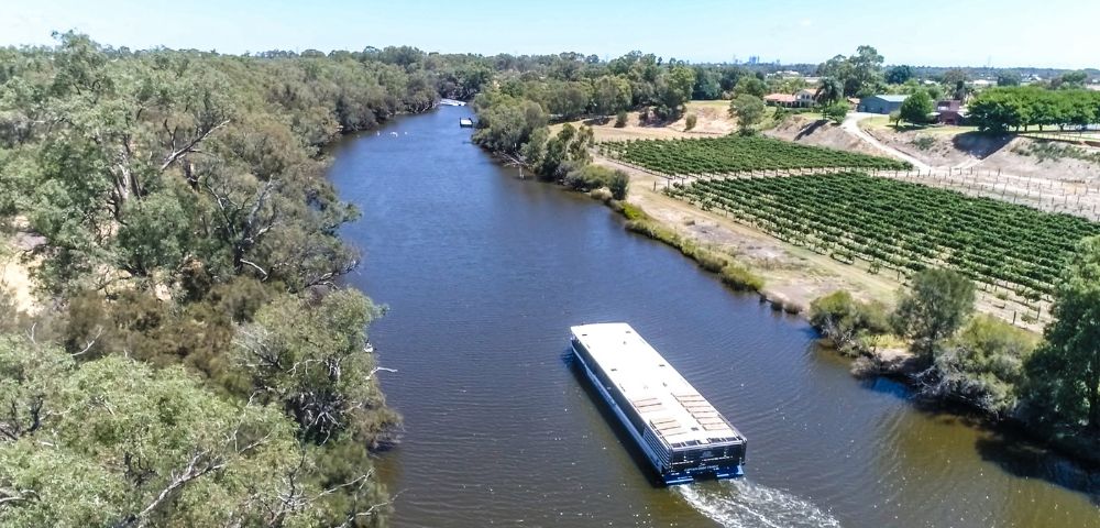 Aerial view of a river with a boat traveling through. Lush trees line the riverbanks, and a vineyard is visible on the right. The scene is peaceful and sunny.