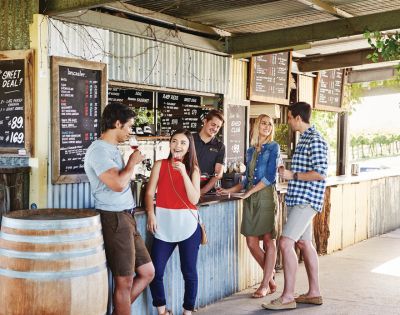 A group of five people at an outdoor cafe counter, enjoying ice cream and chatting. Menu boards and a rustic barrel complete the relaxed, joyful scene.