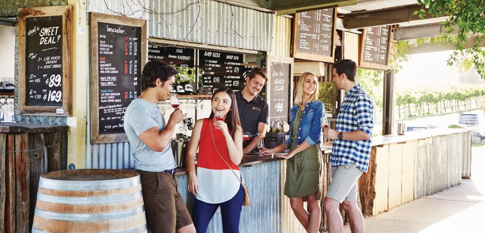 A group of five people at an outdoor cafe counter, enjoying ice cream and chatting. Menu boards and a rustic barrel complete the relaxed, joyful scene.