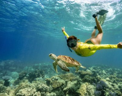 A snorkeler in a yellow rash guard swims underwater alongside a sea turtle above a vibrant coral reef, conveying a sense of adventure and serenity.