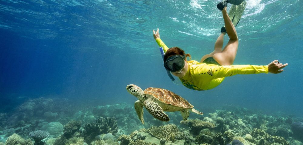 A snorkeler in a yellow rash guard swims underwater alongside a sea turtle above a vibrant coral reef, conveying a sense of adventure and serenity.