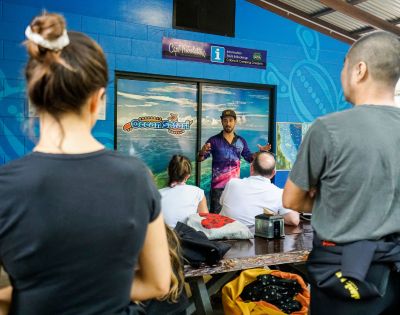 A tour guide passionately speaks to a seated group indoors, against an ocean-themed backdrop. The setting is lively and educational.