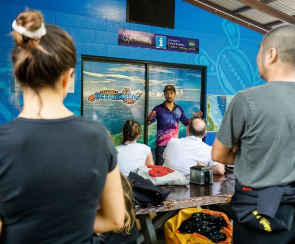 A tour guide passionately speaks to a seated group indoors, against an ocean-themed backdrop. The setting is lively and educational.