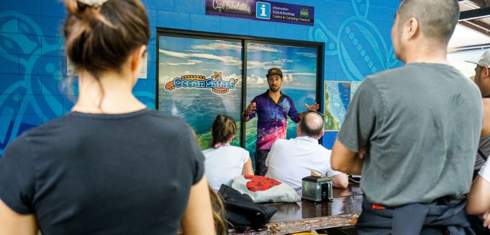 A tour guide passionately speaks to a seated group indoors, against an ocean-themed backdrop. The setting is lively and educational.