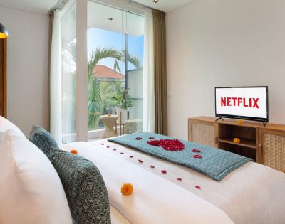 Bright bedroom with cozy, white bed adorned with rose petals and teal accents, a TV showing 