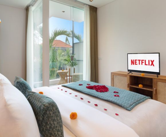 Bright bedroom with cozy, white bed adorned with rose petals and teal accents, a TV showing 