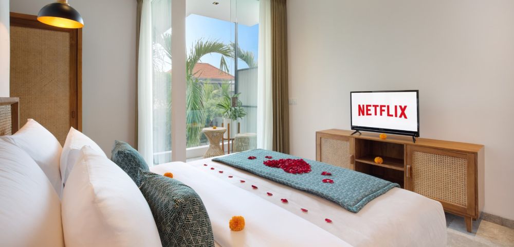 Bright bedroom with cozy, white bed adorned with rose petals and teal accents, a TV showing 