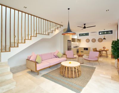 Modern living room with a pink sofa set and pastel cushions. A wooden staircase leads upstairs. Open kitchen and dining area with natural light and decor.