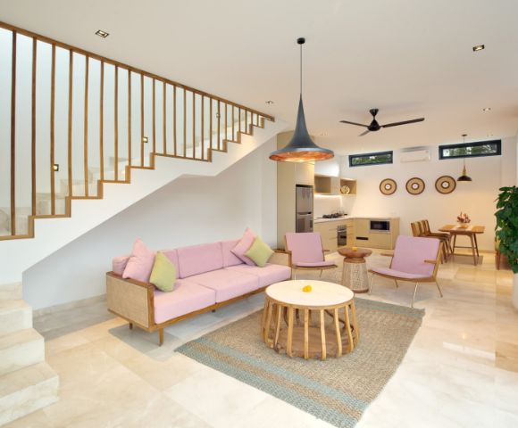 Modern living room with a pink sofa set and pastel cushions. A wooden staircase leads upstairs. Open kitchen and dining area with natural light and decor.