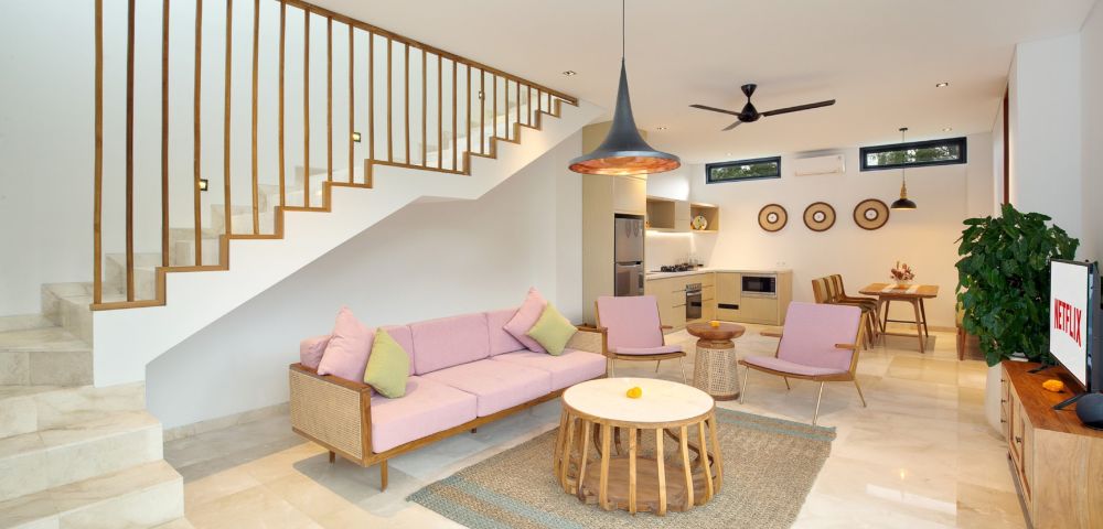 Modern living room with a pink sofa set and pastel cushions. A wooden staircase leads upstairs. Open kitchen and dining area with natural light and decor.