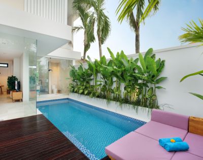 Modern patio with a small blue-tiled pool, surrounded by lush green plants and palm trees. Lounge area features a pink cushion and blue towels, exuding relaxation.