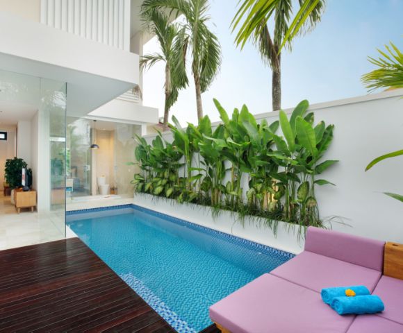 Modern patio with a small blue-tiled pool, surrounded by lush green plants and palm trees. Lounge area features a pink cushion and blue towels, exuding relaxation.
