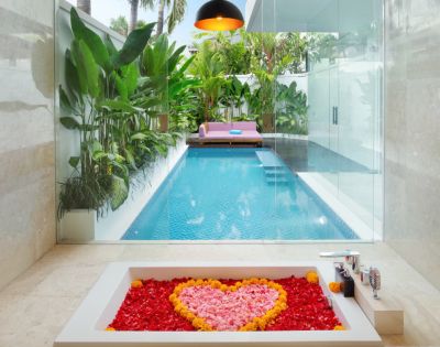 Luxurious bathroom view of a pool through glass. Tub filled with red petals and a heart of pink and yellow. Tropical plants and pink sofa outside. Cozy ambiance.