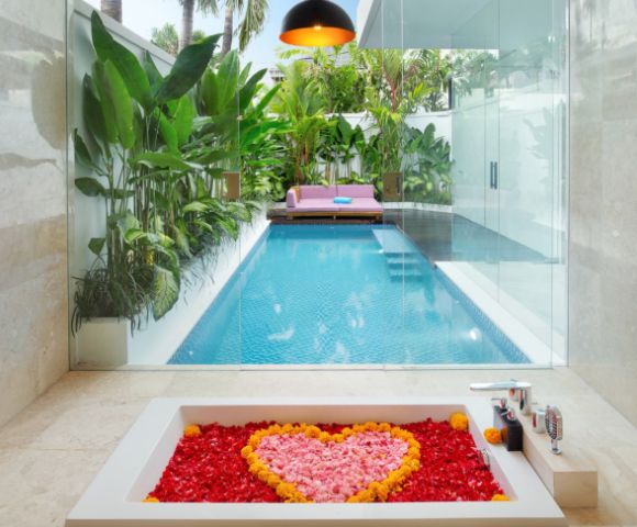 Luxurious bathroom view of a pool through glass. Tub filled with red petals and a heart of pink and yellow. Tropical plants and pink sofa outside. Cozy ambiance.